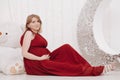 Young beautiful stylish pregnant woman in red long evening dress on white background of child room Royalty Free Stock Photo
