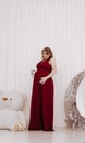 Young beautiful stylish pregnant woman in red long evening dress on white background of child room Royalty Free Stock Photo