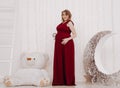 Young beautiful stylish pregnant woman in red long evening dress on white background of child room Royalty Free Stock Photo