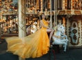 Young beautiful stylish woman sits astride a toy horse, rides a carousel. Long yellow bright dress fluttering in motion