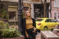 Young beautiful stylish woman in trendy clothes standing on empty city street and looking at camera