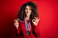 Young beautiful succesful woman with curly hair and piercing winning medals doing ok sign with fingers, excellent symbol