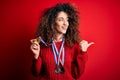 Young beautiful succesful woman with curly hair and piercing winning medals pointing and showing with thumb up to the side with