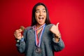 Young beautiful successful asian woman wearing medals standing over red background pointing and showing with thumb up to the side Royalty Free Stock Photo