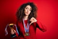 Young beautiful successful woman with curly hair and piercing holding trophy wearing medals with angry face, negative sign showing