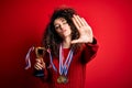 Young beautiful successful woman with curly hair and piercing holding trophy wearing medals with open hand doing stop sign with