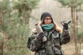 Young beautiful surprised blond woman in camouflage outfit and green scarf posing with thermos in the forest, pointing at