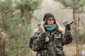 Young beautiful surprised blond woman in camouflage outfit and green scarf posing with thermos in the forest, pointing at