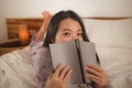 Young beautiful and sweet Asian Korean woman in bed reading book novel feeling happy and playful or studying at home wearing cute Royalty Free Stock Photo