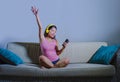 Young beautiful and sweet Asian Korean woman listening to music relaxed and happy sitting at home sofa couch holding mobile phone