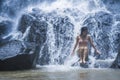 Young beautiful and sweet Asian woman in bikini getting body wet under stream of natural amazing waterfall sitting on rock feeling