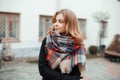 Young beautiful sweet woman in a trendy winter warm coat with a stylish woolen checkered scarf stands on a warm autumn day