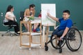 Young and beautiful teacher teaching cute down syndrome girl while disabled boy on wheelchair painting and drawing on paper in