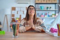 Young beautiful teacher woman wearing sweater and glasses sitting on desk at kindergarten begging and praying with hands together Royalty Free Stock Photo