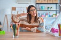 Young beautiful teacher woman wearing sweater and glasses sitting on desk at kindergarten gesturing with hands showing big and Royalty Free Stock Photo