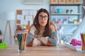 Young beautiful teacher woman wearing sweater and glasses sitting on desk at kindergarten looking sleepy and tired, exhausted for Royalty Free Stock Photo