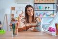 Young beautiful teacher woman wearing sweater and glasses sitting on desk at kindergarten smiling and confident gesturing with Royalty Free Stock Photo