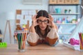 Young beautiful teacher woman wearing sweater and glasses sitting on desk at kindergarten Trying to open eyes with fingers, sleepy Royalty Free Stock Photo