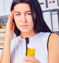 Young beautiful tired sick woman sitting in workplace in office holding bottle with pills. Female feeling bad at work.