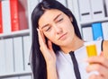 Young beautiful tired sick woman sitting in workplace in office holding bottle with pills. Female feeling bad at work.