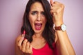 Young beautiful woman applying red lipstick over pink isolated background annoyed and frustrated shouting with anger, crazy and Royalty Free Stock Photo