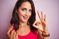 Young beautiful woman applying red lipstick over pink isolated background doing ok sign with fingers, excellent symbol Royalty Free Stock Photo