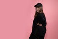 A young beautiful woman in black casual clothes and sports baseball cap stands with back half-sided to the camera and looks into