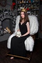 woman in a black dress with a golden crown on her head and a skull in her hands is sitting in a rocking chair near the fireplace.