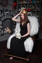 woman in a black dress with a golden crown on her head and a skull in her hands is sitting in a rocking chair near the fireplace.