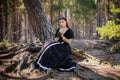 Young, beautiful woman in a black medieval dress with a steel rose in her hands, sitting in the woods on the roots of a tree. Attr