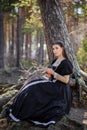 Young, beautiful woman in a black medieval dress with a steel rose in her hands, sitting in the woods on the roots of a tree. Attr