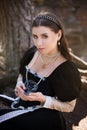 Young, beautiful woman in a black medieval dress with a steel rose in her hands, sitting in the woods on the roots of a tree. Attr