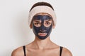 Young beautiful woman in black mud mask for face doing cosmetic procedures, looks sad, posing against white wall, wearing hair Royalty Free Stock Photo