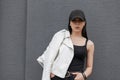 Young beautiful woman in a black T-shirt in a white stylish leather jacket in a trendy black cap poses near a gray building Royalty Free Stock Photo