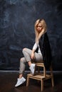 Young beautiful woman with blonde hair in a stylish black shirt, white sneakers and torn jeans sitting on a wooden chair on a dar Royalty Free Stock Photo