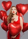 Young beautiful woman blowing kiss and holding red balloons. Model girl with red lips makeup wearing red dress on holiday party Royalty Free Stock Photo