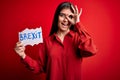 Young beautiful woman with blue eyes holding paper with brexit message over red background with happy face smiling doing ok sign Royalty Free Stock Photo