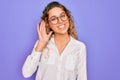 Young beautiful woman with blue eyes wearing casual shirt and glasses over purple background smiling with hand over ear listening Royalty Free Stock Photo