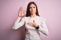 Young beautiful woman with blue eyes wearing casual white t-shirt over pink background Swearing with hand on chest and open palm,