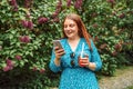 Young beautiful woman in bright blue dress holding plastic cold tea cup and looking at smartphone while walking at city Royalty Free Stock Photo