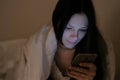 Young beautiful woman brunette looks at social networks in her mobile phone in bed before sleeping and smiles. Royalty Free Stock Photo