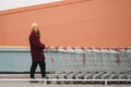 Young beautiful woman in a burgundy coat and yellow hat walks on the roof of supermarket parking lot and takes trolley. Royalty Free Stock Photo