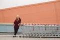 Young beautiful woman in a burgundy coat and yellow hat walks on the roof of supermarket parking lot and takes trolley. Royalty Free Stock Photo