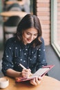 Young beautiful woman in the cafe near the window, thinking and writing something. Royalty Free Stock Photo