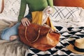 Young beautiful woman in casual clothes sitting on bed at home and putting her yellow autumn scarf in leather handbag Royalty Free Stock Photo