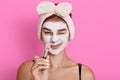 Young beautiful woman with clay facial mask on her face holding brush in hands after doing beauty treatment, making funny face Royalty Free Stock Photo