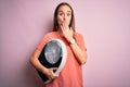 Young beautiful woman controlling weight holding weighting machine over pink background cover mouth with hand shocked with shame