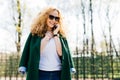 Young beautiful woman with curly blonde hair wearing sunglasses and green jacket talking on her cellphone, smiling while standing Royalty Free Stock Photo