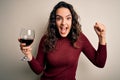 Young beautiful woman with curly hair drinking glass of red wine over white background screaming proud and celebrating victory and Royalty Free Stock Photo