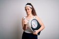 Young beautiful woman with curly hair holding weighing machine over white background doing money gesture with hands, asking for